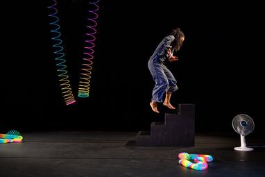 Vergrößerte Ansicht von Eine Frau schwebt über einer vierstufigen Treppe. Rechts neben der Treppe steht ein Ventilator. Auf dem Boden liegen bunte Spiralen und eine bunte Spirale hängt in der Luft.