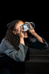 Vergrößerte Ansicht von Eine Frau in einer grauen Jacke biegt eine silberne Spirale und schaut durch sie hindurch wie durch ein Fernglas.