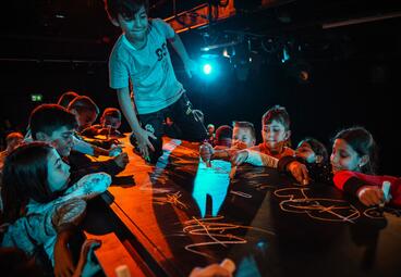 Vergrößerte Ansicht von Eine Gruppe Kinder steht um einen tisch, welcher zu hoch für sie ist. Sie kritzeln mit Kreide auf der Oberfläche herum. Ein Kind läuft auf Knien über den Tisch.