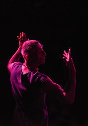 Vergrößerte Ansicht von Uwe Topppmann hebt seine Arme und schaut hinter sich fragend ins Publikum. Von links wird er mit einem pinken Licht beleuchtet.