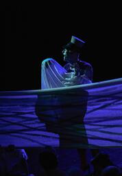 Vergrößerte Ansicht von Lila Beleuchtung. Uwe Toppmann trägt eine Schaffnermütze auf dem Kopf und redet mit seiner hand, welche sich unter einem weißen Laken befindet.