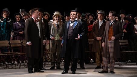 Vergrößerte Ansicht von Theaterszene aus Pagliacci mit mehreren Darstellern in historischen Kostümen, darunter Frack, Zylinder und Westen, auf einer Bühne mit Stuhlreihen im Hintergrund.