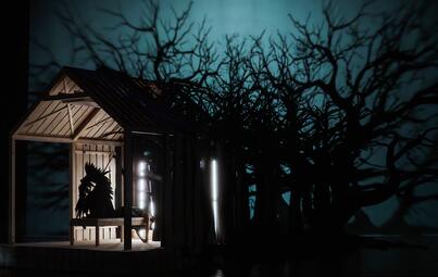 Vergrößerte Ansicht von Dunkle Bühne mit Holzhaus, darin sitzende Figur mit Krone, umgeben von Schatten von kahlen Bäumen, Szene aus der Oper Lohengrin.