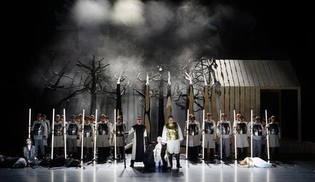 Vergrößerte Ansicht von Bühnenbild der Oper Lohengrin mit kahlen Bäumen, Chor in weißen Kostümen, zwei Figuren in der Mitte, eine kniet, eine trägt goldene Rüstung, rechts Holzhaus, Nebel im Hintergrund.