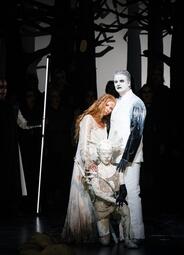 Vergrößerte Ansicht von Drei Personen in Kostümen auf einer Bühne mit dunklen Baum-Silhouetten im Hintergrund, eine Person kniet, eine andere trägt ein langes, helles Kleid.