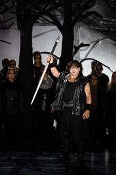 Vergrößerte Ansicht von Sänger in schwarzer Lederweste und schwarzer Hose hält Schwert mit Kreuzgriff erhoben, Hintergrund mit dunklen Baum-Silhouetten und weiteren Personen in dunkler Kleidung.