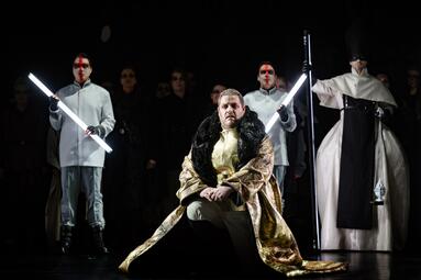 Vergrößerte Ansicht von Szene aus der Oper Lohengrin mit einer Person in goldenem Gewand und schwarzem Pelz, umgeben von Personen in weißen Kostümen, die lange Leuchtstäbe halten.