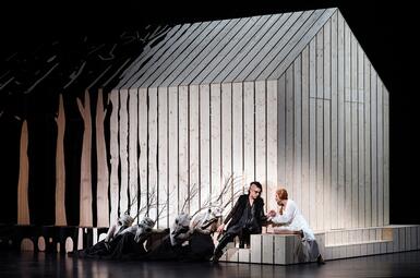 Vergrößerte Ansicht von Bühnenbild mit hellem Holzhaus und stilisierten Bäumen, Schauspieler in dunkler und weißer Kostümierung, sitzend und kniend, Szene aus der Oper Lohengrin.