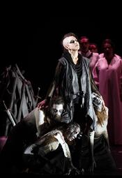 Vergrößerte Ansicht von Dramatische Szene aus der Oper Lohengrin mit einer stehenden Person in dunklem, zerrissenem Kostüm, umgeben von knienden Personen in hellen Gewändern, im Hintergrund weitere Figuren in weißen Kleidern.