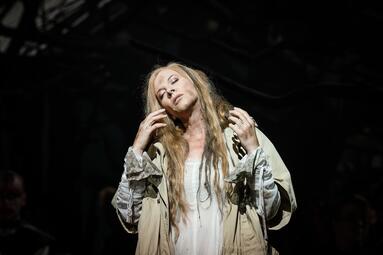 Vergrößerte Ansicht von Person mit langen, offenen Haaren trägt ein helles, locker sitzendes Kleid und eine beige Jacke, hebt die Hände zum Kopf, dunkler Hintergrund.