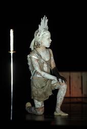 Vergrößerte Ansicht von Kostümierte Person kniet mit weißer Körperbemalung und Federkopfschmuck auf dunkler Bühne, neben einem langen Schwert in Scheide.