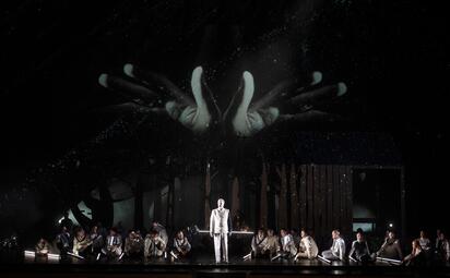 Vergrößerte Ansicht von Bühnenbild der Oper Lohengrin mit großem, leuchtendem Handmotiv im Hintergrund und Ensemble in historischen Kostümen, zentrale Figur in hellem Anzug.