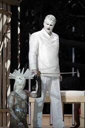 Vergrößerte Ansicht von Zwei Personen in weißer Kleidung, eine steht mit einem Schwert, die andere kniet mit einem Helm, Szene aus der Oper Lohengrin.