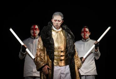 Vergrößerte Ansicht von Person in goldfarbenem Gewand mit schwarzem Pelzkragen, flankiert von zwei Personen in weißen Hemden, die lange weiße Stäbe halten.