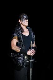 Vergrößerte Ansicht von Schauspieler in schwarzer, glänzender Weste und schwarzem Unterhemd hält Schwert mit beiden Händen vor dem Körper, Szene aus der Oper Lohengrin.