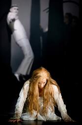 Vergrößerte Ansicht von Schauspielerin mit langen roten Haaren in weißem Kostüm kniet auf dem Boden, im Hintergrund eine weitere Person in weißem Anzug, Szene aus der Oper Lohengrin.