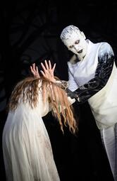 Vergrößerte Ansicht von Zwei Personen in weißen Kostümen auf dunkler Bühne, eine mit langen roten Haaren verbeugt sich, die andere trägt schwarze Handschuhe und hält die Arme der ersten Person.
