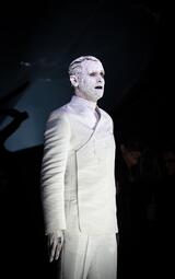 Vergrößerte Ansicht von Person in weißem, zweireihigem Anzug mit hochgeschlossenem Hemd, steht vor dunklem Hintergrund, Szene aus der Oper Lohengrin.