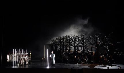 Vergrößerte Ansicht von Bühnenbild der Oper Lohengrin mit dunklem Wald aus kahlen Bäumen, Figuren in weißen Kostümen und einer Person mit leuchtendem Stab im Vordergrund.