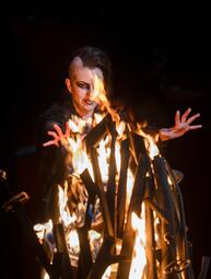 Vergrößerte Ansicht von Person mit seitlich rasiertem Haar und langem Deckhaar streckt die Hände über ein brennendes Holzfeuer in dunkler Umgebung.