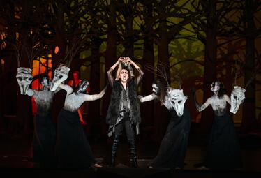 Vergrößerte Ansicht von Szene aus der Oper Lohengrin mit fünf Personen, vier tragen schwarze Kleider und weiße Tiermasken, eine Person in schwarzer Kleidung mit Fellweste hebt die Hände über den Kopf.