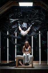 Vergrößerte Ansicht von Szene aus der Oper Lohengrin mit einer sitzenden Frau in weißem Kleid und einem hinter ihr stehenden Mann mit schwarzem Flügelkostüm und silberner Kopfbedeckung.