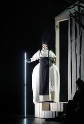Vergrößerte Ansicht von Person in weißem, voluminösem Kostüm mit schwarzem Latz steht auf hölzernem Podest neben einem beleuchteten Stab, Szene aus der Oper Lohengrin.