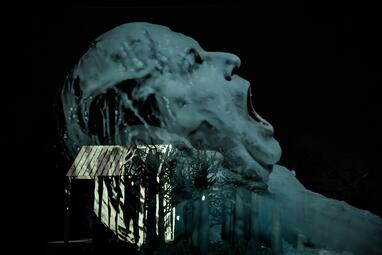 Vergrößerte Ansicht von Große, blaue Skulptur eines schreienden Kopfes vor dunklem Hintergrund, davor schwarze Silhouetten von Bäumen und einem kleinen Haus.