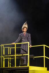 Vergrößerte Ansicht von Person mit hochgesteckter, spitz zulaufender Frisur trägt einen schwarz-weiß gestreiften Anzug mit roter Krawatte und steht auf einem gelben Geländer.