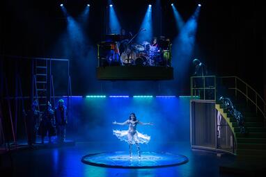 Vergrößerte Ansicht von Bühnenbild der Produktion »Slippery Slope« am Nationaltheater Mannheim mit einer Tänzerin in der Mitte, einer schwebenden Band mit Schlagzeug und Bass, sowie mehreren Personen auf der Bühne und einer Treppe rechts.