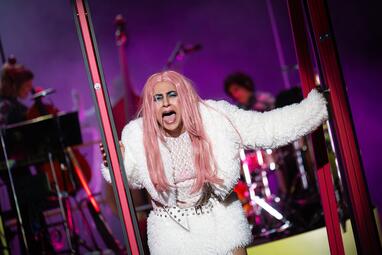 Vergrößerte Ansicht von Schauspielerin mit langen rosa Haaren trägt weißes, flauschiges Kostüm und steht mit ausgestreckten Armen zwischen Glaswänden auf der Bühne.