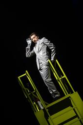 Vergrößerte Ansicht von Person in silbernem Anzug steht auf gelbem Gerüst vor schwarzem Hintergrund, eine Hand an den Kopf gelegt.