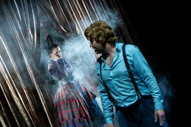 Vergrößerte Ansicht von Zwei Schauspieler auf der Bühne, eine trägt ein blaues Hemd mit Hosenträgern, die andere ein kariertes Kleid mit Puffärmeln, umgeben von Nebel und Vorhängen.