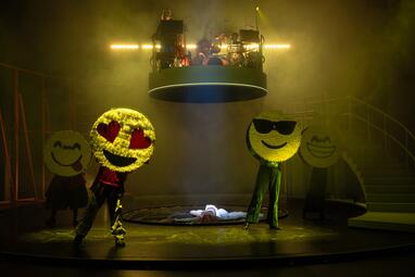 Vergrößerte Ansicht von Bühnenbild der Produktion »Slippery Slope« mit vier Personen in großen Emoji-Kostümen, darunter Herzaugen- und Sonnenbrillen-Emoji, und einer Person liegend auf der Bühne unter einer runden Plattform mit Musikern.