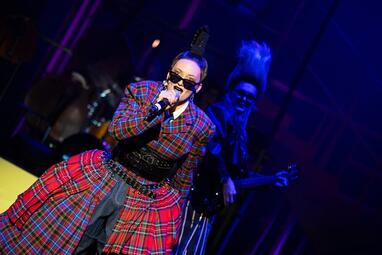 Vergrößerte Ansicht von Sängerin in rotem, kariertem Kostüm mit schwarzem Korsett und Federhaarschmuck auf der Bühne, im Hintergrund ein Musiker mit E-Gitarre.