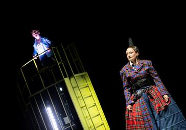 Vergrößerte Ansicht von Schauspielerin in kariertem Kostüm mit hohem Hut und breitem Gürtel steht rechts, links Person in blauer Jacke auf gelbem Metallgerüst.