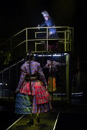 Vergrößerte Ansicht von Schauspielerin in kariertem, mehrlagigem Rock und Rollschuhen steht vor einer erhöhten Plattform mit Geländer, auf der eine weitere Person in gestreiftem Mantel steht.