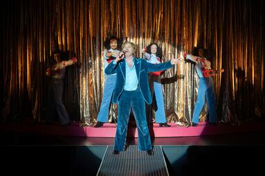 Vergrößerte Ansicht von Fünf Personen auf Bühne vor goldglänzendem Vorhang, vorn ein Mann im blauen Samtanzug mit ausgestreckten Armen, dahinter vier Personen in blauen Overalls mit roten Gitarren.