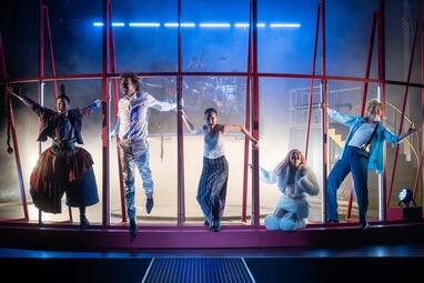 Vergrößerte Ansicht von Fünf Schauspieler in verschiedenen Kostümen stehen und sitzen an einem roten Gitterrahmen auf der Bühne.