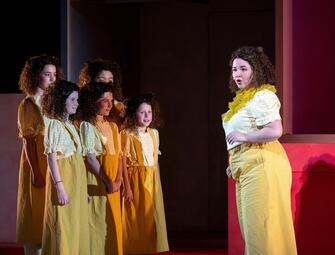 Vergrößerte Ansicht von Fünf Personen in gelben Kleidern stehen links, eine Person in gelbem Oberteil und Hose rechts vor rotem Hintergrund.