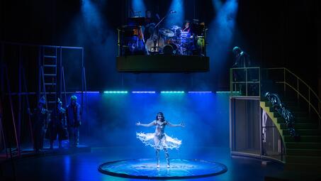 Vergrößerte Ansicht von Bühnenbild der Produktion »Slippery Slope« am Nationaltheater Mannheim mit einer Tänzerin in der Mitte, einer schwebenden Band mit Schlagzeug und Bass, sowie mehreren Personen auf der Bühne und einer Treppe rechts.