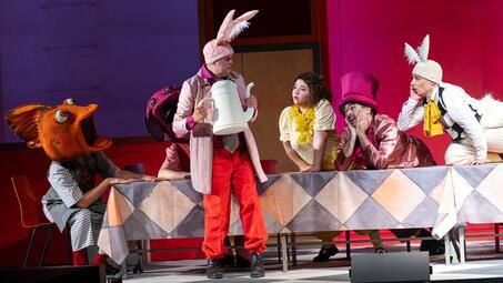 Vergrößerte Ansicht von Fünf Schauspieler in Kostümen aus der Theaterinszenierung von Alice im Wunderland sitzen und stehen um einen mit einem Rautenmuster bedeckten Tisch, darunter zwei mit Hasenohren und ein mit einem großen roten Hut.