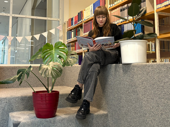 Vergrößerte Ansicht von Eine junge Frau sitzt auf der Lesetreppe in der Zentralbibliothek Mannheim und liest in dem Buch &quot;Der Weihnachtspulli&quot;.