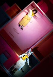 Vergrößerte Ansicht von Person in gelber Hose und weißem Oberteil liegt auf einer pinkfarbenen Wand, darunter steht eine Person mit weißem Hasenkostüm und gelbem Halstuch in einem Raum mit roten Wänden.