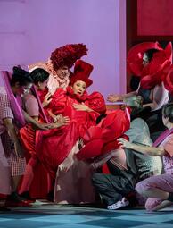 Vergrößerte Ansicht von Mehrere Personen in bunten Kostümen, darunter zwei in auffälligen roten Kleidern mit großen roten Hüten, umgeben eine sitzende Person in einem roten Kleid.