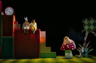 Vergrößerte Ansicht von Zwei Personen in bunten Kostümen sitzen auf einer mehrstufigen, farbigen Bühne, rechts eine große Pilz- und Pflanzen-Dekoration.