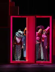 Vergrößerte Ansicht von Mehrere Personen in bunten Kostümen stehen in einem rot beleuchteten, rechteckigen Rahmen auf einer Bühne.