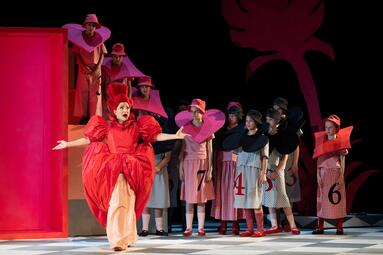 Vergrößerte Ansicht von Eine Frau in einem roten, voluminösen Kleid mit ausgestreckten Armen steht vor einer Gruppe von Personen in karierten Kostümen mit großen, farbigen Kragen und Zahlen auf den Kleidern, auf einer Bühne mit rotem Hintergrund.
