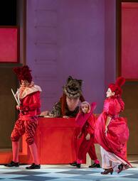 Vergrößerte Ansicht von Drei Personen in roten Kostümen und ein als Katze verkleideter Darsteller auf einer Bühne mit roten und braunen Kulissenteilen.