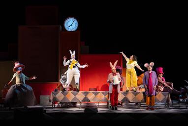 Vergrößerte Ansicht von Bühnenbild mit mehreren Schauspielern in bunten Kostümen, darunter ein weißer Hase mit gelber Weste und eine Frau in gelber Hose, vor einer großen Uhr und roten Kulissen.